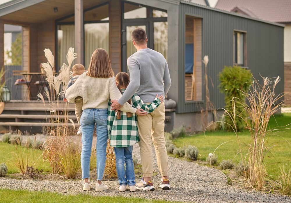 Happy family. View from back of man woman with child and girl standing together hugging and admiring their cozy home on fine autumn day